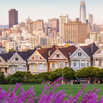 Pastel evening view of San Francisco’s Painted Ladies with twilight hues, city skyline in the distance, and flowers in the foreground.