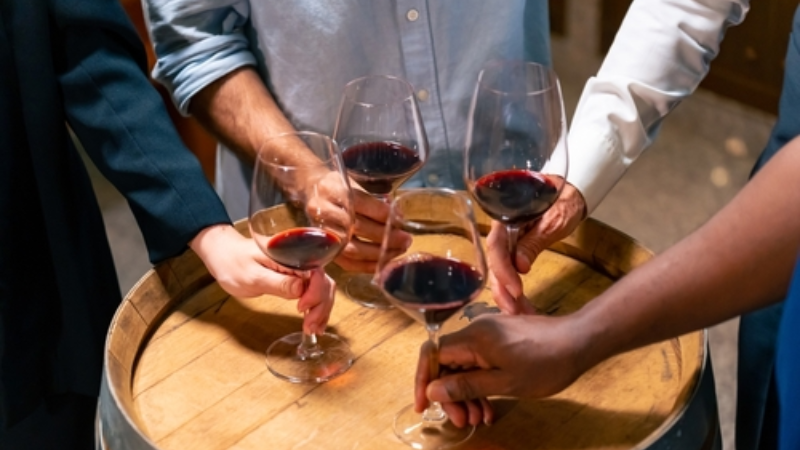 Group of professionals tasting red wine in glasses over wooden barrels, participating in a formal work event in the wine cellar.