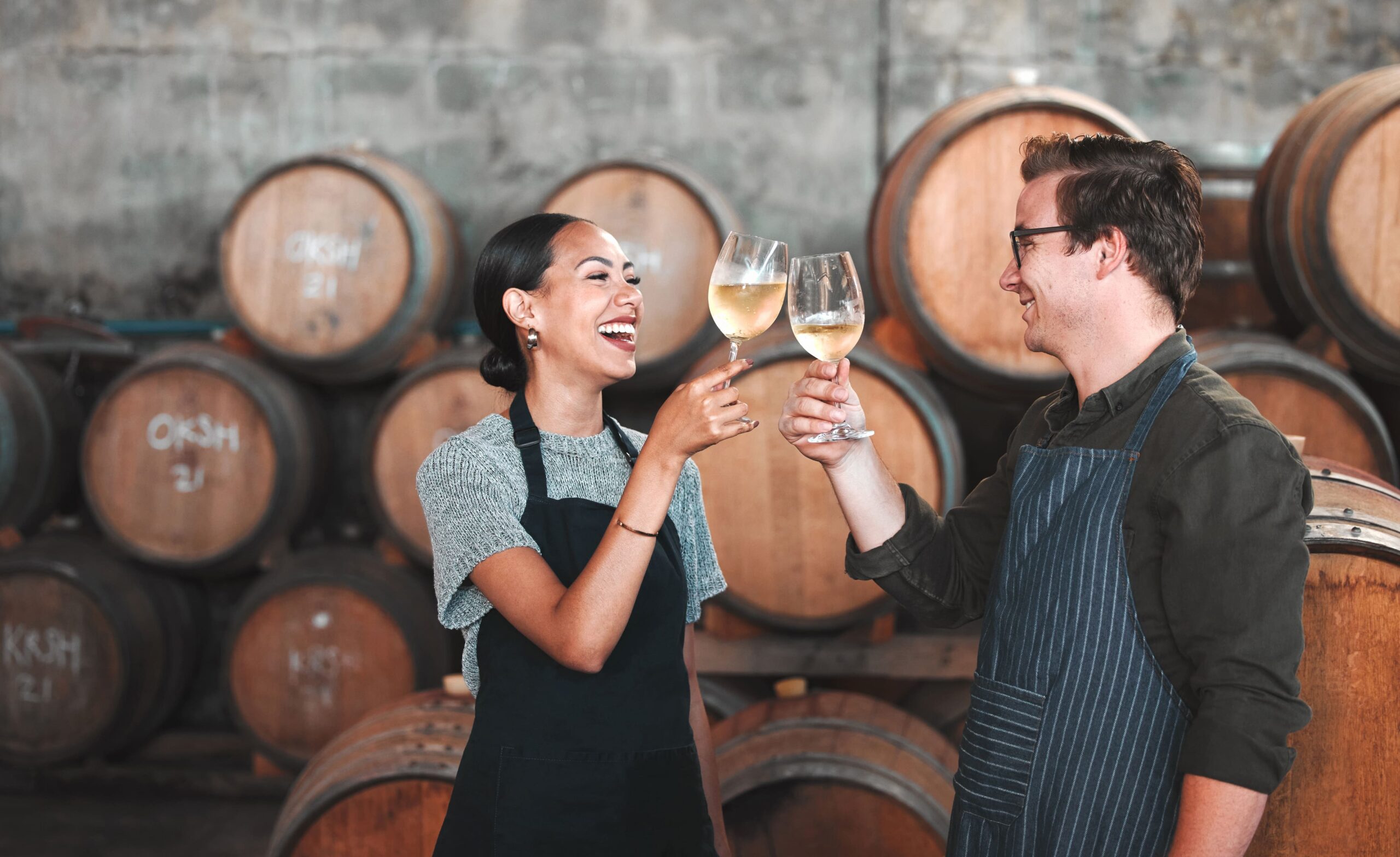 A couple toasts to each other with glasses of wine in a wine cellar surrounded by barrels.