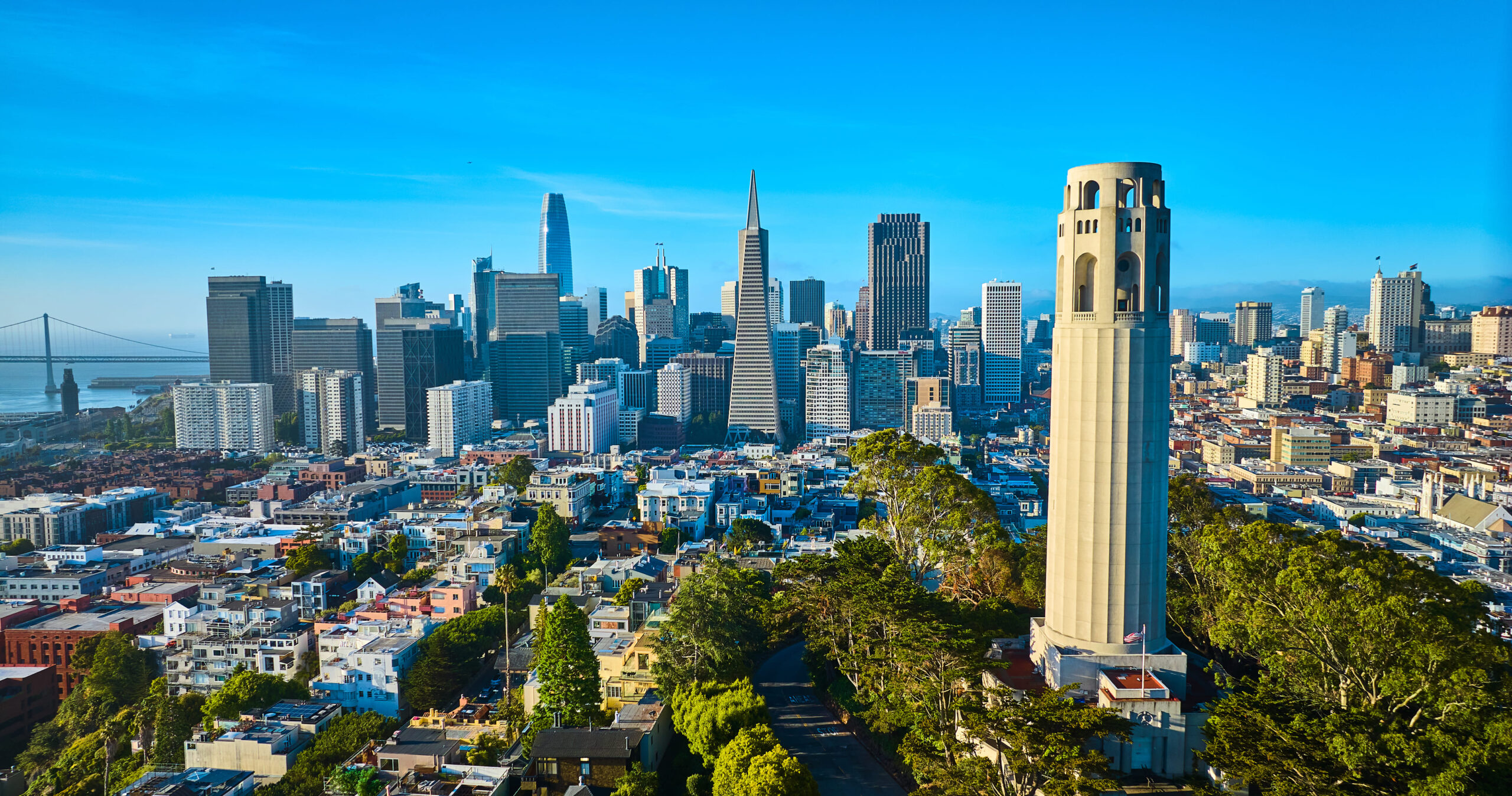 Aerial view of Coit Tower in the late afternoon with downtown San Francisco skyscrapers and the Bay Bridge in the distance.