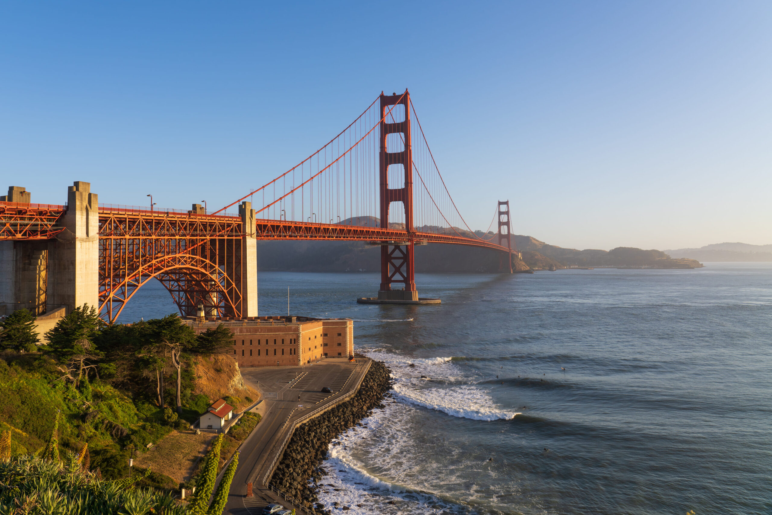 Amazing view of the Golden Gate bridge baked in warm orange morning sunlight, showcasing the mist from the ocean and the deep blue sky.