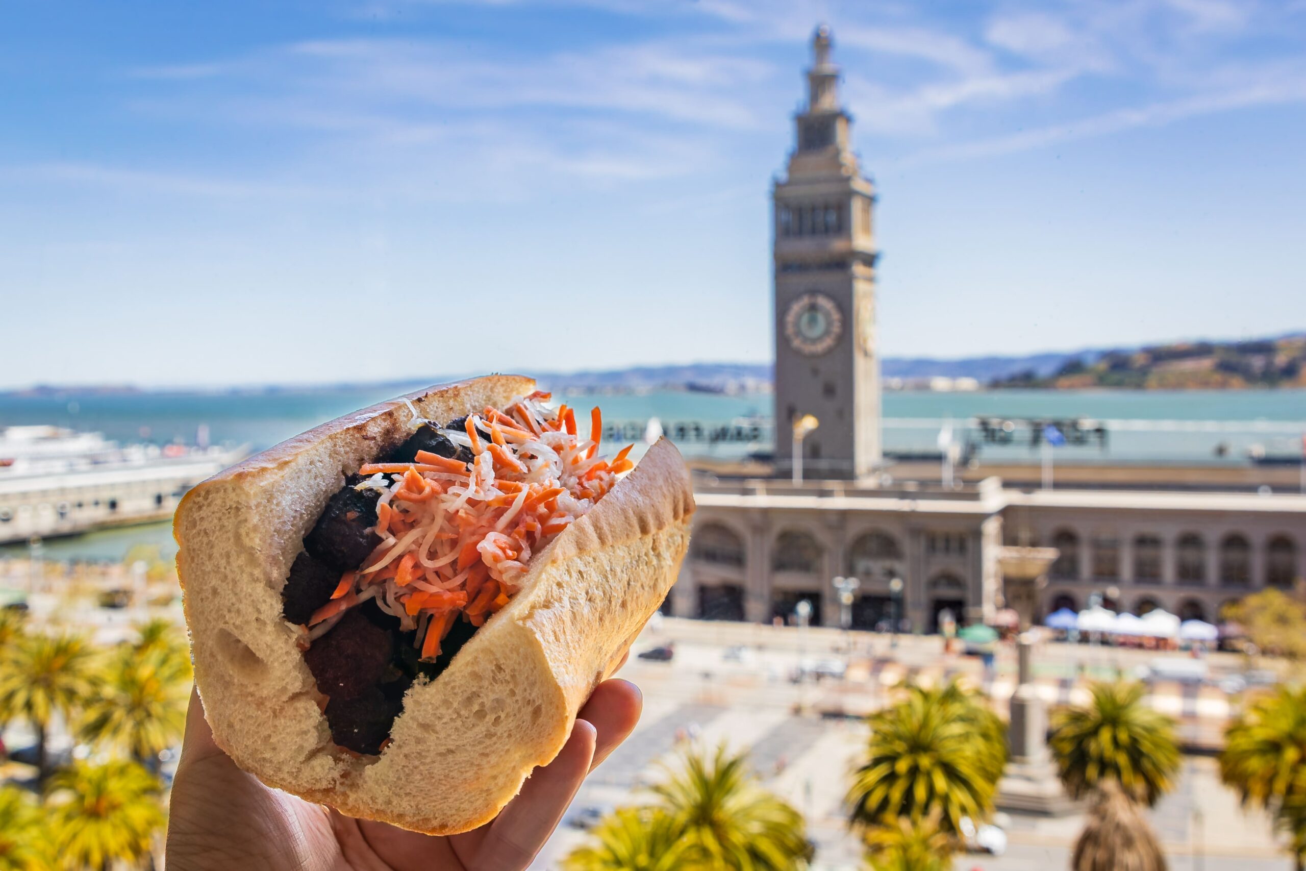 Someone holding up a banh mi sandwich against the backdrop of the San Francisco Ferry Building in San Francisco, CA.